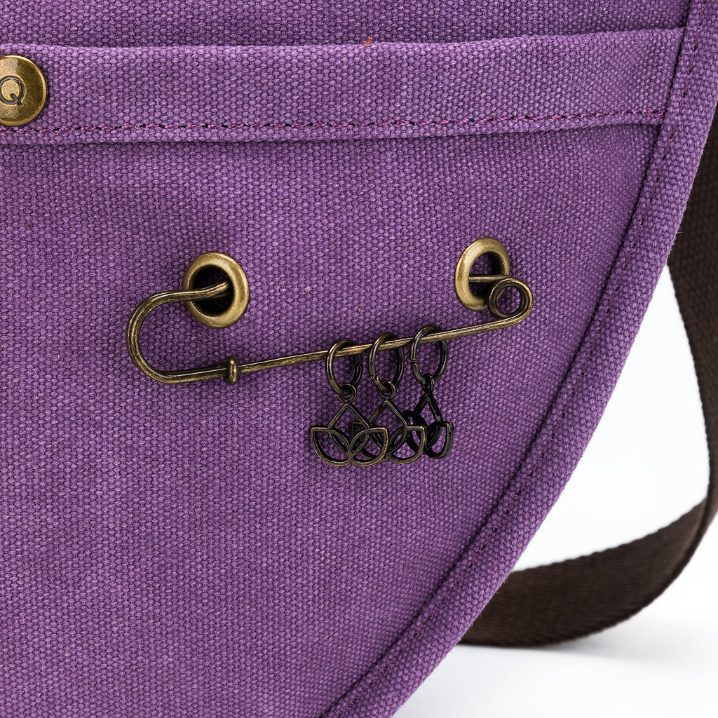 Close-up of a brass safety pin-style loop on a purple della Q bag, holding several black stitch markers shaped like abstract leaves or petals.