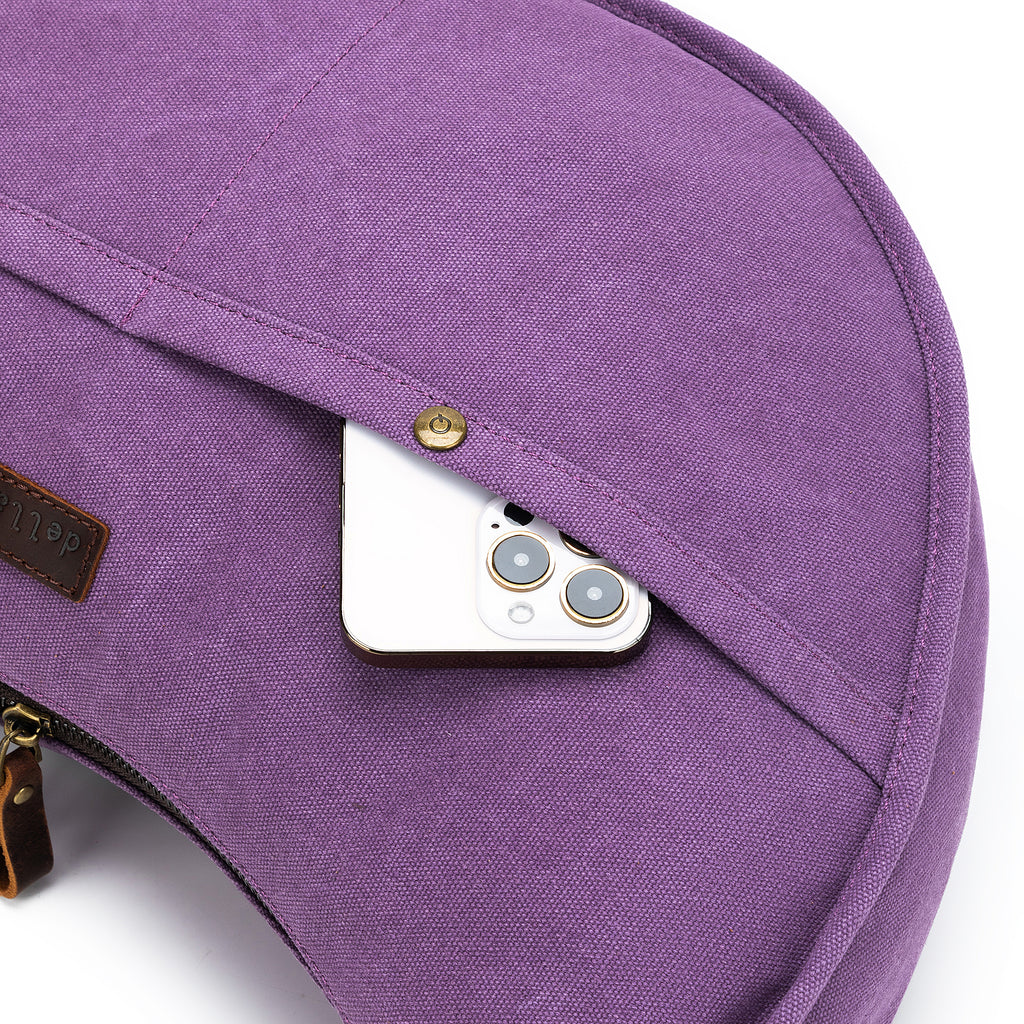 A white smartphone is partially tucked into the outer pocket of a purple della Q bag. Brass snaps and a leather logo patch are visible.