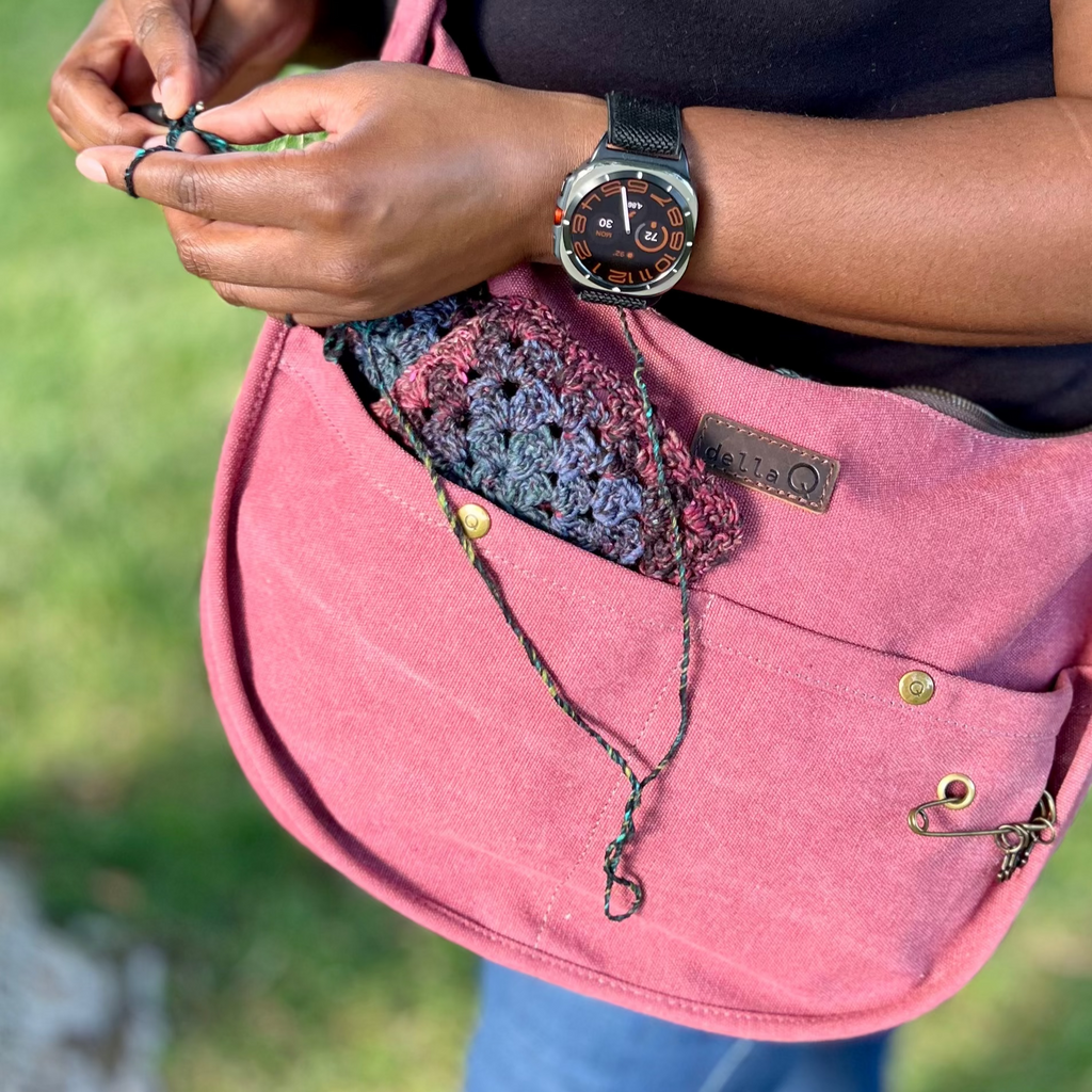 A person stands outdoors wearing a black shirt and jeans, holding a crochet project. A maroon adella Q bag with a granny square project inside hangs from their shoulder.