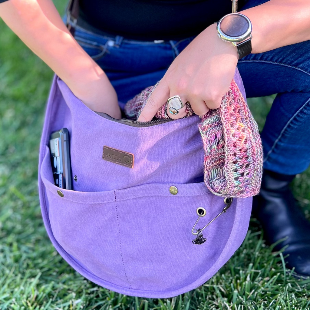 A person wearing a black top and jeans reaches into a purple della Q bag while holding a multicolored project. The bag rests on green grass and the person wears rings and a smartwatch.
