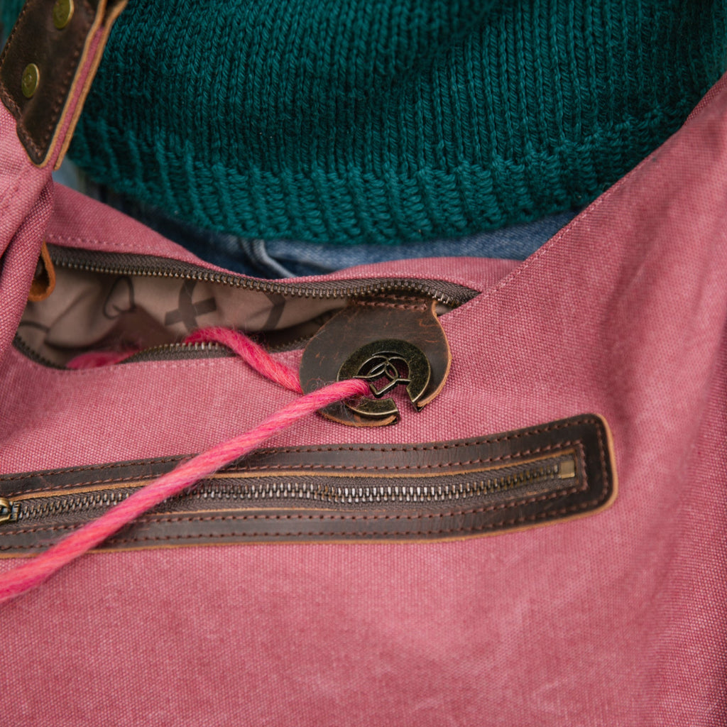 A red della Q bag sits against a person’s torso, with a ball of pink yarn being fed cleanly through a yarn feeder cut-out. The bag is partially zipped with knitting visible inside.